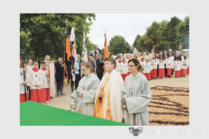 Ungewohntes Bild hinter Kaplan Robert Ginter, Zelebrant am vierten Altar &ndash; dem langen, hei&szlig;en und vor allem trockenen Sommer geschuldet, gab es in diesem Jahr nicht gen&uuml;gend Bl&uuml;tenbl&auml;tter, um einen Blumenteppich zu streuen. Also musste man sich mit Holzschnitzeln behelfen.
(Fotos: A. No&eacute;)