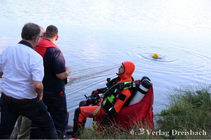 Der Sicherungstaucher wartet am Ufer in voller Ausrüstung, während sein Kollege im Okrifteler Baggerseee taucht. Der Signalmann hat die rote Leine des Tauchenden in den Händen und beobachtet das Geschehen auf und im Wasser.
