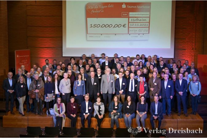 Oliver Klink (links), Vorstandsvorsitzender der Taunus Sparkasse, Landrat Michael Cyriax (vorne rechts sitzend), Verwaltungsratsvorsitzender, und Raimund Becker (2. v.r. auf der Bühne), Mitglied des Vorstands, zusammen mit Vertreterinnen und Vertretern der 113 Vereine im Landratsamt Hofheim.