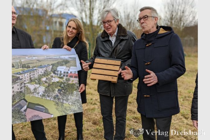Hubert Nopper (Mitte) zeigt symbolisch ein Stück Holz, das auf die in modularer Holzbauweise entstehenden Wohngebäude hinweisen soll.
