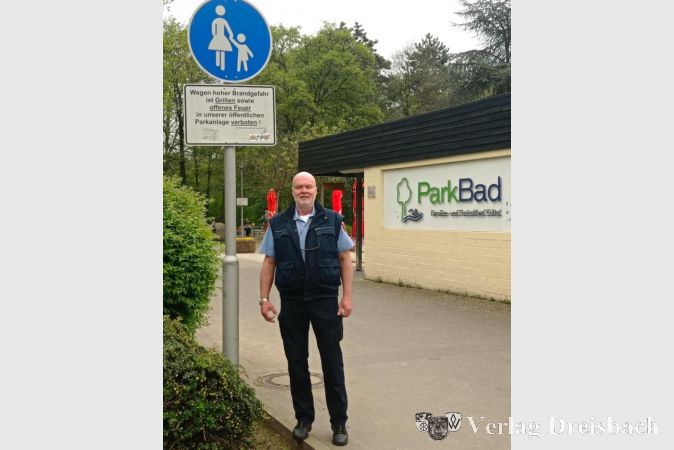 Hilfspolizist Stefan Lörsch neben einem blauen Gehweg-Schild am Eingang zum Freizeitpark (Höhe Parkbad). Das Schild besagt: „Der Weg darf nur als Gehweg von Fußgängern genutzt werden. Für alle anderen Verkehrsteilnehmer ist der Weg verboten.“ Fahrradfahrer/innen müssen absteigen und ihr Rad schieben.