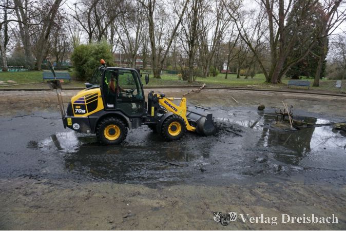 Radlader bei der Säuberung des Weihers. Auch wenn die Fontäne Sauerstoff ins Wasser befördert, bilden sich nach und nach Algen im Wasser; altes Laub, Äste und Dreck sammeln sich vor allem in Herbst und Winter dort. Einen natürlichen Zu- und Abfluss gibt es nicht. Damit das beliebte Ausflugsziel, ursprünglich als Motorboot-Fahrfläche angelegt, nicht zum Sumpf wird, muss das Wasser abgelassen, Dreck aus dem Becken geschaufelt und alles ausgespritzt werden. Dann kann das frische Wasser einlaufen.